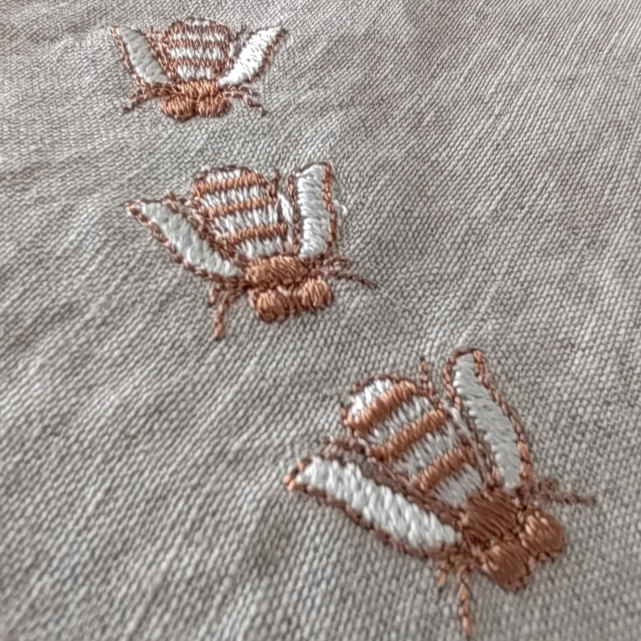 Close-up of a light gray textured fabric with three embroidered butterflies in dusty rose and white thread. MI RE LA