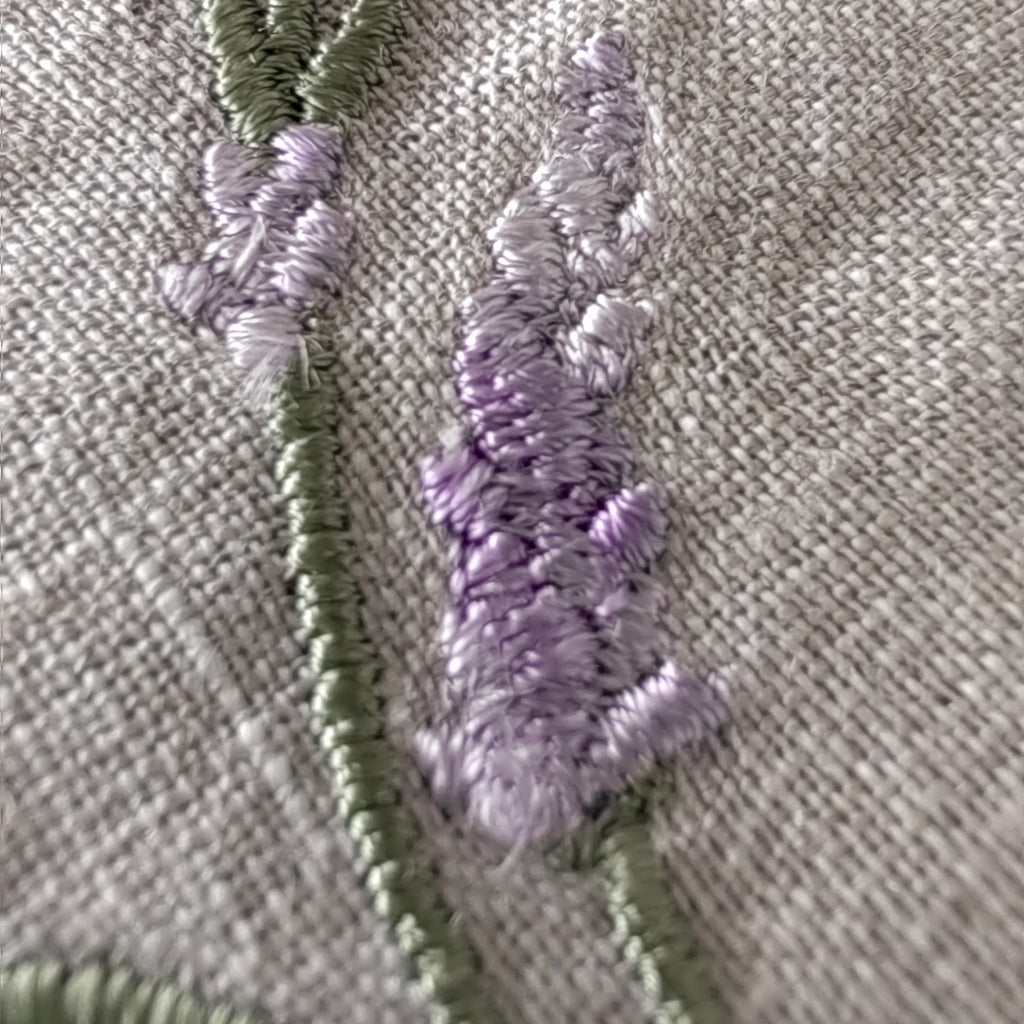 Close-up of embroidered lavender stems in light purple and sage green on a textured beige fabric background. MI RE LA