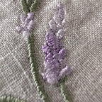 Close-up of embroidered lavender stems in light purple and sage green on a textured beige fabric background. MI RE LA