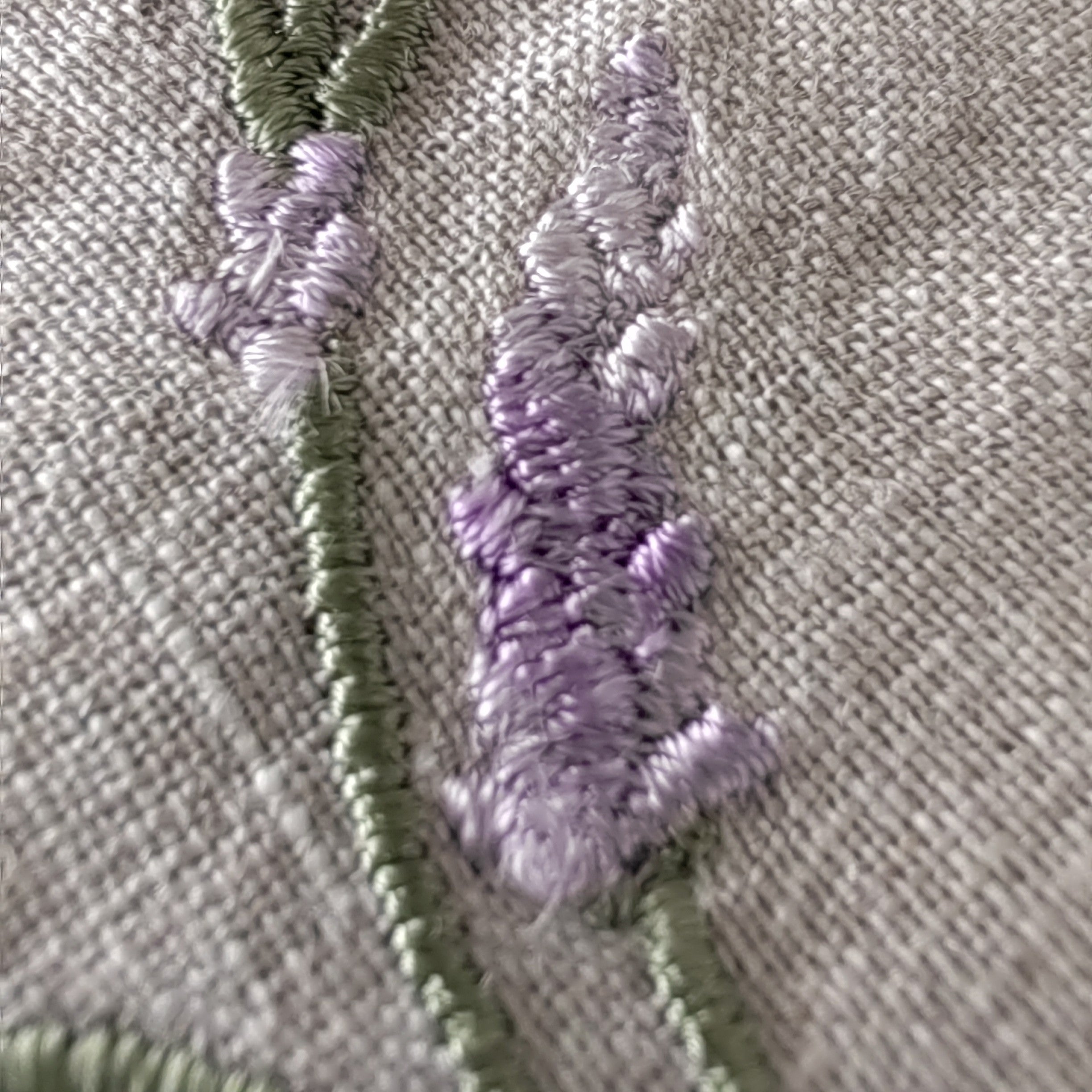 Close-up of embroidered lavender stems in light purple and sage green on a textured beige fabric background. MI RE LA