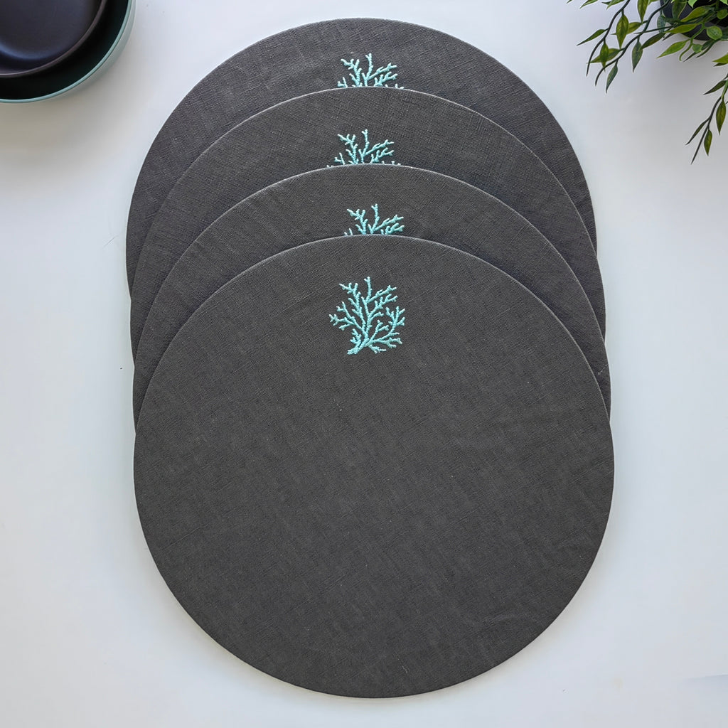Four round dark gray placemats with a small light blue coral embroidery, arranged in a fan shape on a white surface.