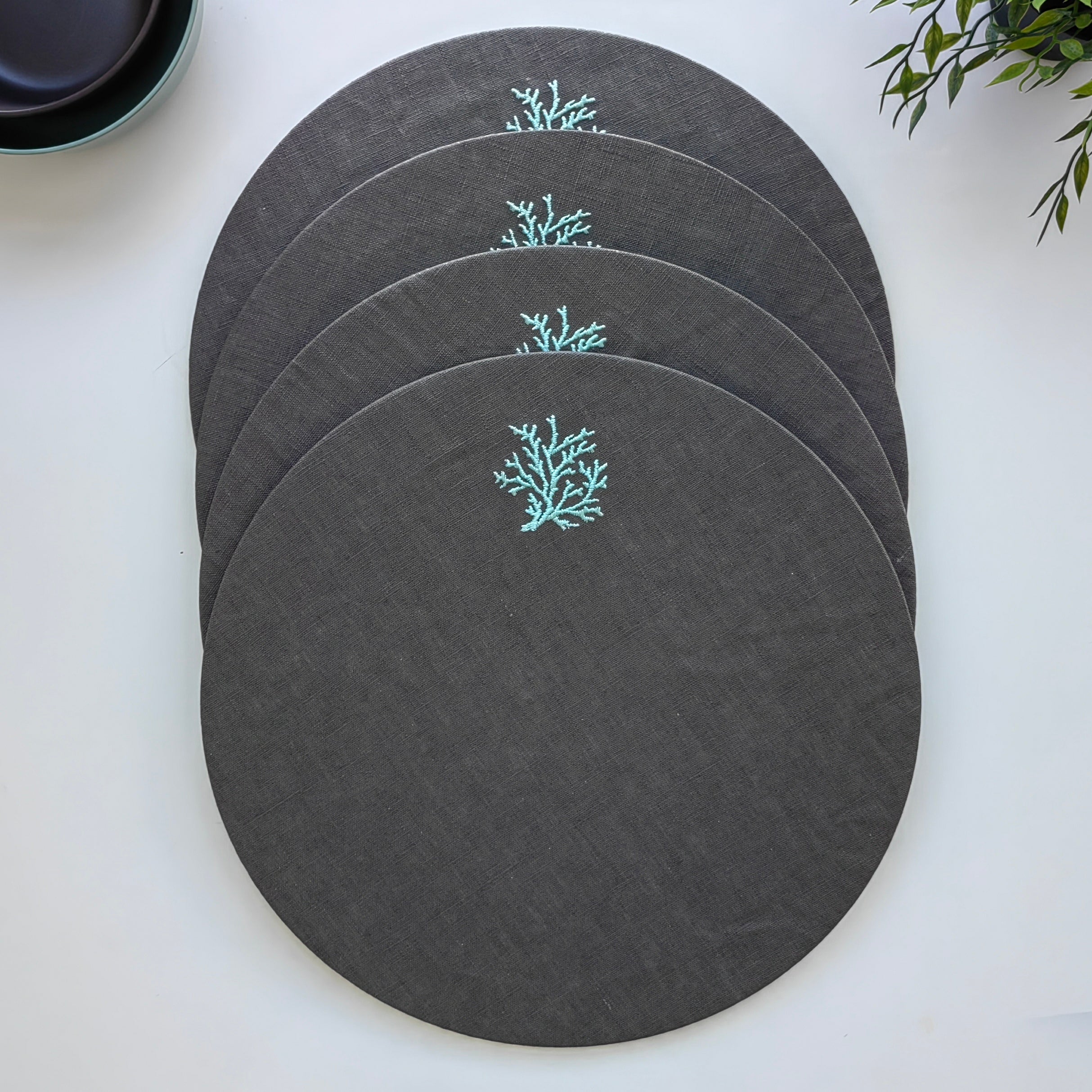 Four round dark gray placemats with a small light blue coral embroidery, arranged in a fan shape on a white surface.