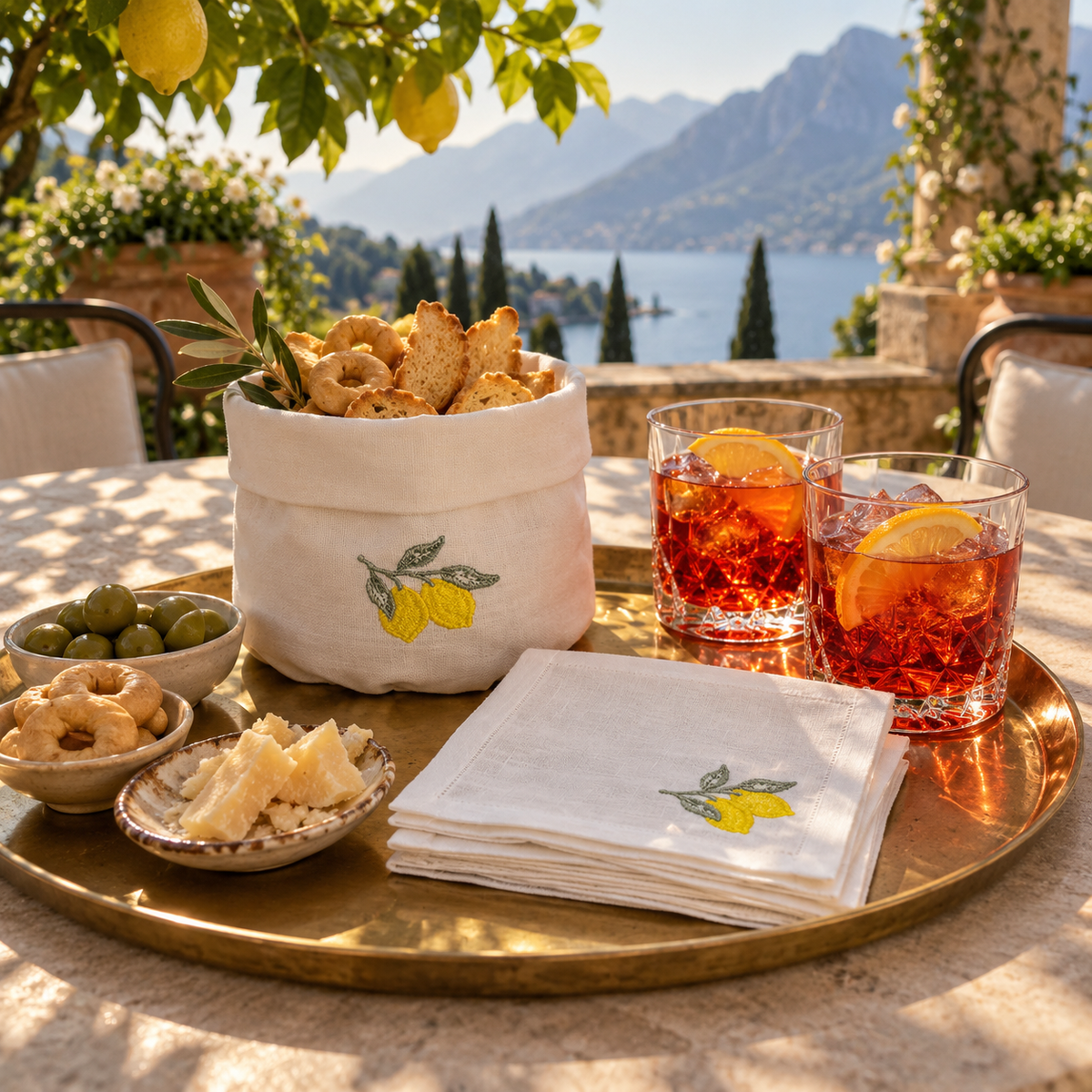 A styled outdoor dining setup on a round brass tray, featuring a white linen napkin and bread basket embroidered with yellow lemons, alongside glasses of red cocktail with lemon slices and small bowls of olives and cheese, set against a scenic lake and mountain background.