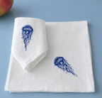 Two folded white napkins with blue jellyfish embroidery, displayed on a light blue background with a small apple partially visible in the top-left corner. MI RE LA