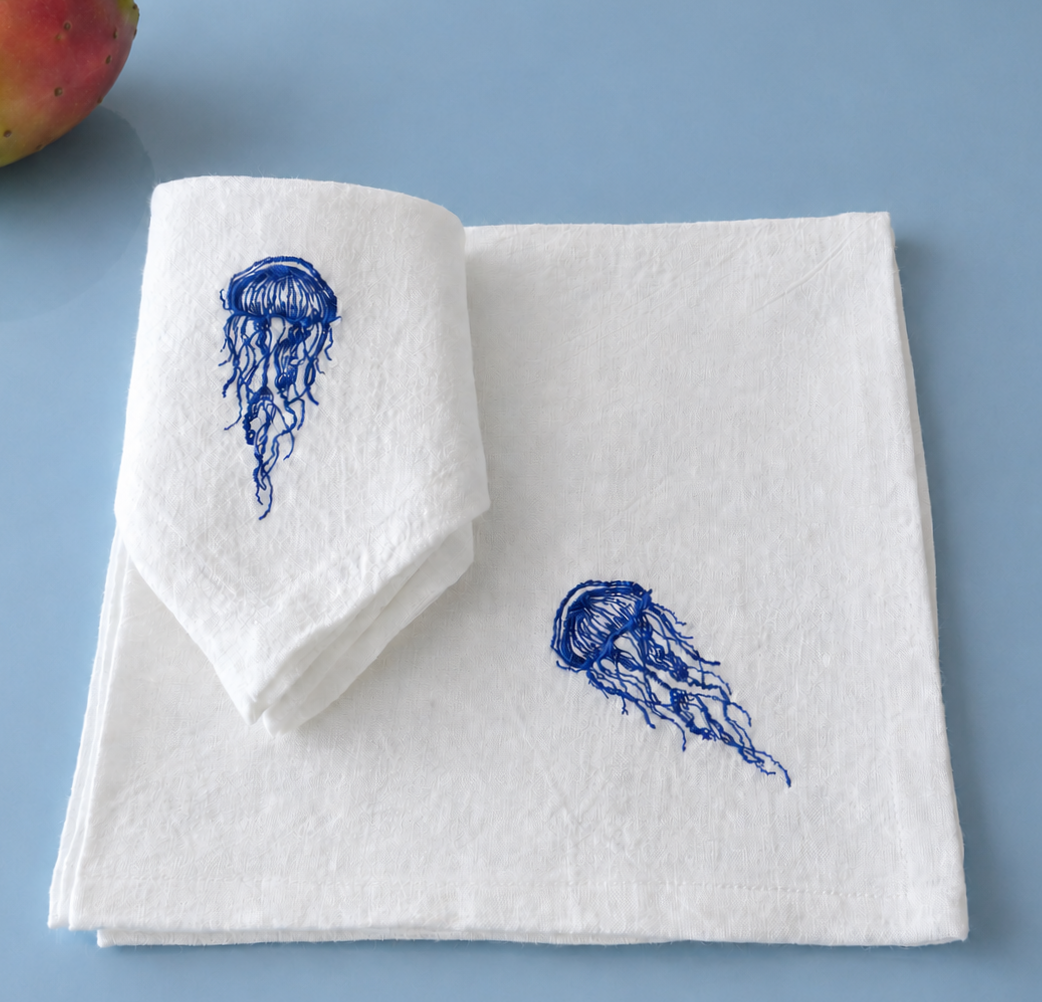 Two folded white napkins with blue jellyfish embroidery, displayed on a light blue background with a small apple partially visible in the top-left corner. MI RE LA