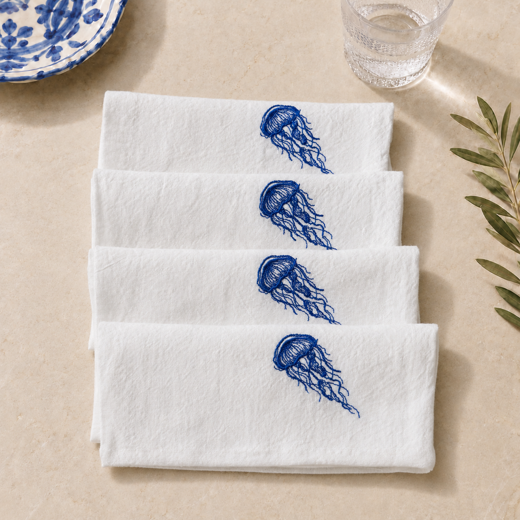 Four white cloth napkins stacked diagonally on a beige surface, each embroidered with a blue jellyfish motif. A blue-and-white ceramic plate edge and a glass are partially visible in the background, along with a leafy sprig.