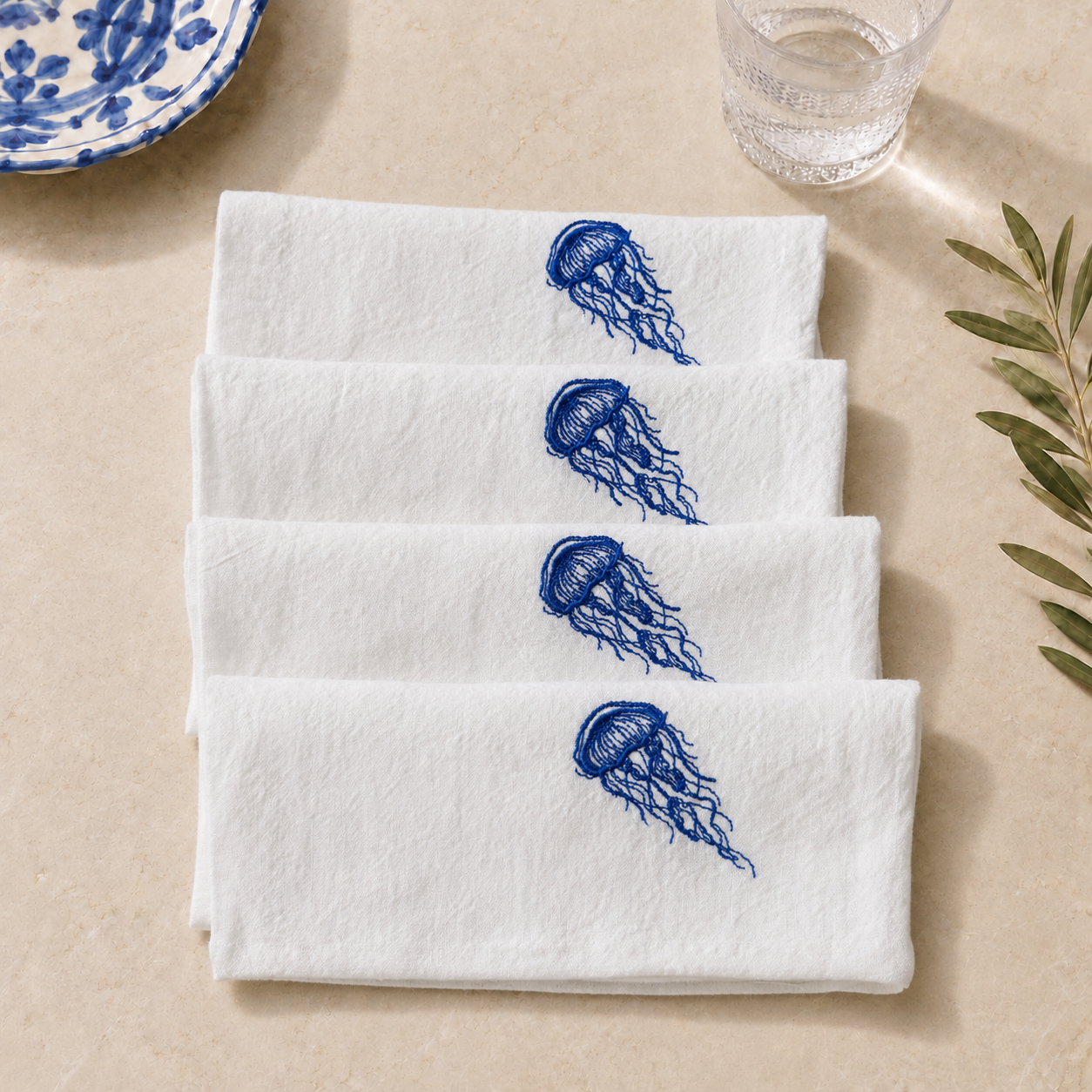 Four white cloth napkins stacked diagonally on a beige surface, each embroidered with a blue jellyfish motif. A blue-and-white ceramic plate edge and a glass are partially visible in the background, along with a leafy sprig.
