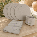 A coordinated linen tableware set displayed outdoors on a wooden surface, including four round placemats, folded napkins, and a small cup or bowl. The items are a light warm beige with embroidered green stems and small purple flowers.
