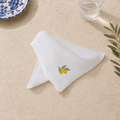 A folded white cotton napkin with a small embroidered yellow lemon and green leaves, styled on a beige textured surface with a glass and a blue patterned plate partially visible. MI RE LA