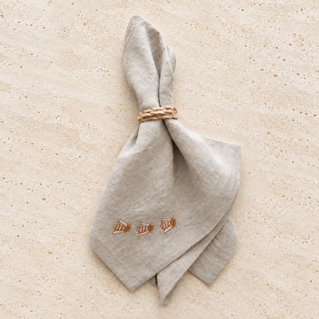 A light beige linen napkin tied with a braided rope napkin ring, featuring embroidered bees near the bottom corner, laid flat on a sandy beige background. MI RE LA