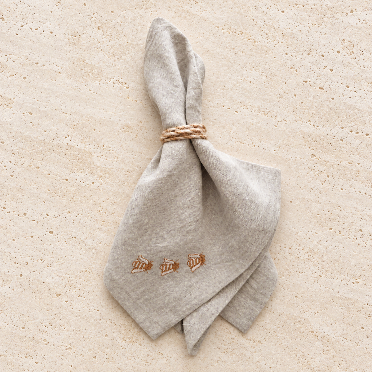 A light beige linen napkin tied with a braided rope napkin ring, featuring embroidered bees near the bottom corner, laid flat on a sandy beige background. MI RE LA