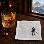 A white linen napkin with a black embroidered skier graphic, displayed on a dark wooden table next to a glass of whiskey with an orange slice, with a mountain window in the background.