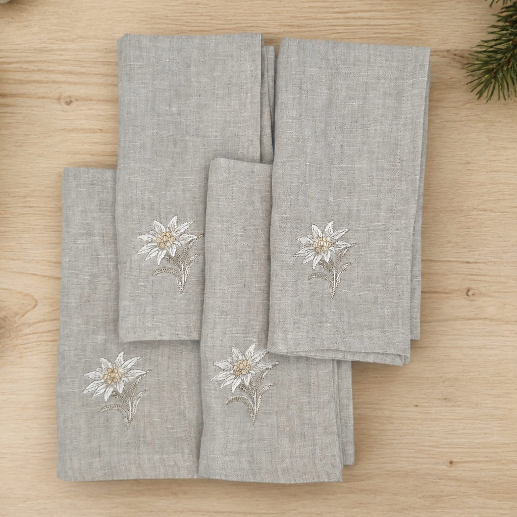 Four light gray linen napkins with embroidered white and gold floral motifs, arranged on a light wood tabletop with pine branches in the corner.