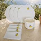 White dinnerware set with embroidered yellow lemons and green leaves, including a stack of plates, folded napkins, and a small bowl, displayed outdoors on a stone surface with greenery and water in the background.
