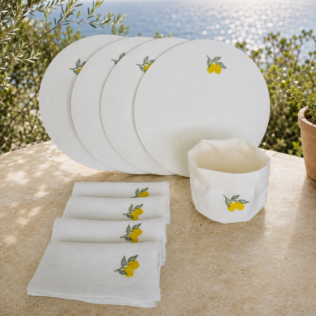 White dinnerware set with embroidered yellow lemons and green leaves, including a stack of plates, folded napkins, and a small bowl, displayed outdoors on a stone surface with greenery and water in the background.