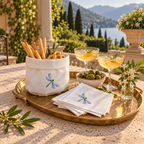 A styled outdoor dining setup on a stone table with a brass tray holding a white napkin holder embroidered with a blue dragonfly, white napkins, breadsticks, olives, and two filled coupe glasses, with a scenic lake and mountains in the background.