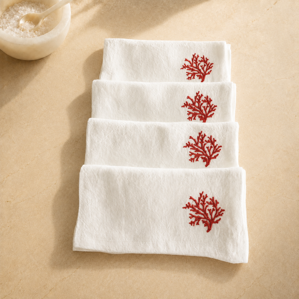 Four folded white towels stacked diagonally on a beige surface, each decorated with a small red coral motif near the top edge. MI RE LA