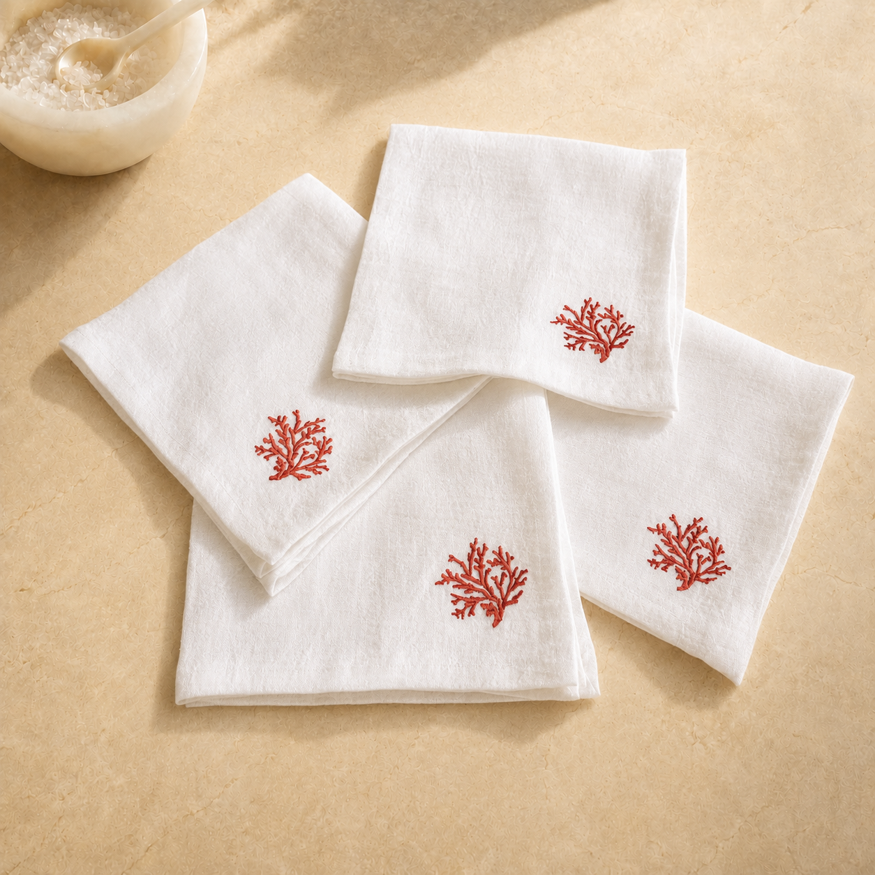 Four white cotton napkins with red coral embroidery, arranged on a beige textured surface with a small bowl in the background. MI RE LA