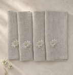 Four folded oatmeal stone linen napkins with white and gold embroidered wildflowers, arranged on a textured beige surface with small dried flowers in the corner.