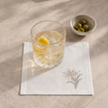 A white linen napkin with a delicate embroidered floral design, styled on a textured beige stone surface. A clear cut-glass tumbler containing a light-colored drink and a lemon wedge sits on the napkin, with a small white bowl of green olives in the background.