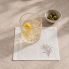 A white linen napkin with a delicate embroidered floral design, styled on a textured beige stone surface. A clear cut-glass tumbler containing a light-colored drink and a lemon wedge sits on the napkin, with a small white bowl of green olives in the background.