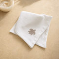 A folded white cotton washcloth with a small brown embroidered sea turtle on the front, displayed on a beige stone surface with a small bowl in the upper left corner.