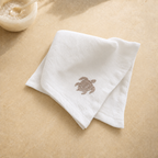 A folded white cotton washcloth with a small brown embroidered sea turtle on the front, displayed on a beige stone surface with a small bowl in the upper left corner.