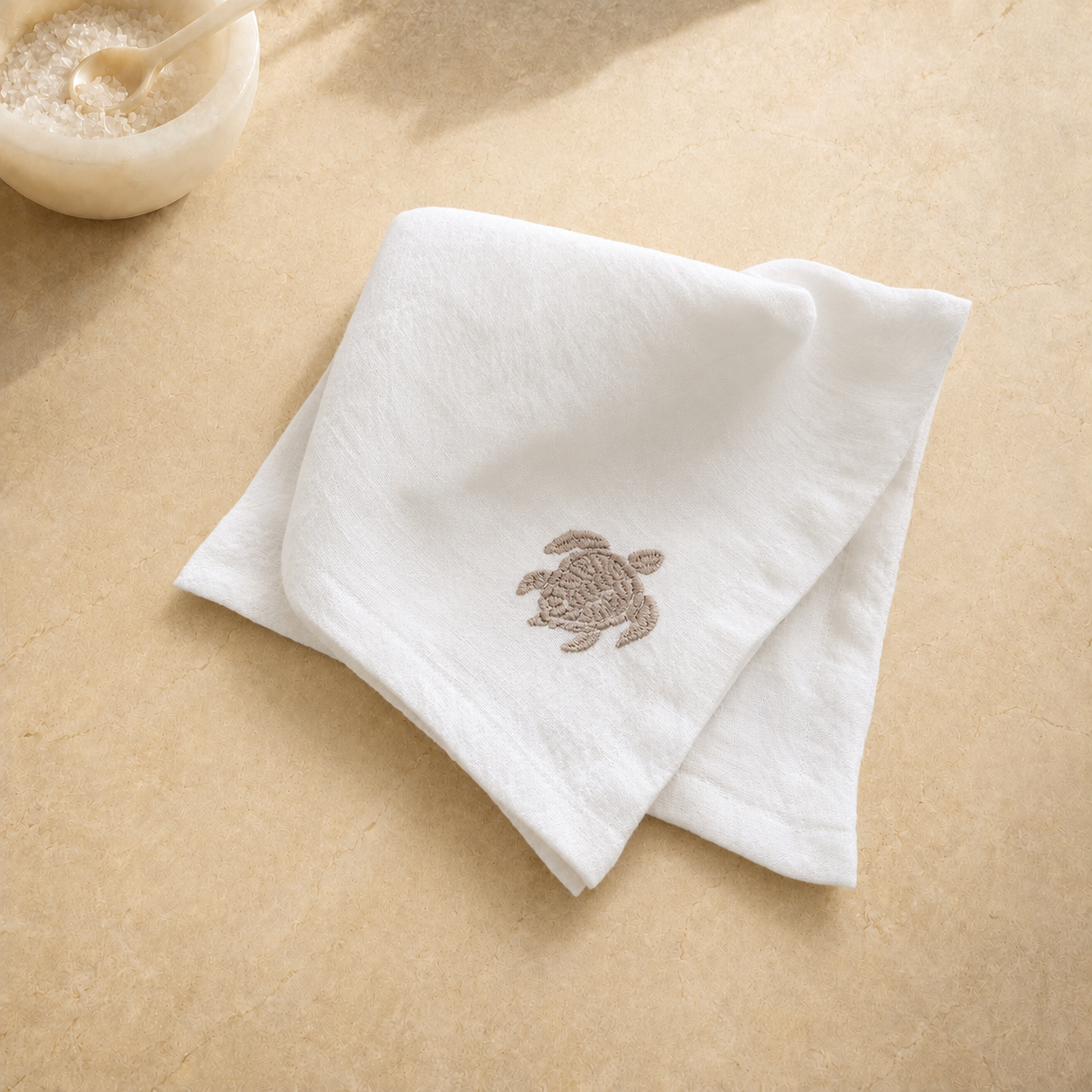 A folded white cotton washcloth with a small brown embroidered sea turtle on the front, displayed on a beige stone surface with a small bowl in the upper left corner.