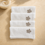 Four folded white towels stacked on a beige surface, each printed with a brown sea turtle design near the top. A small bowl with a spoon is partially visible in the upper left corner. MI RE LA
