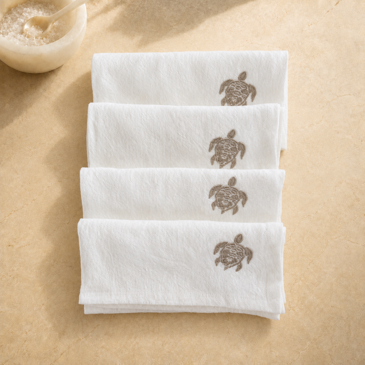 Four folded white towels stacked on a beige surface, each printed with a brown sea turtle design near the top. A small bowl with a spoon is partially visible in the upper left corner. MI RE LA