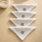 Four white folded cotton napkins with embroidered sea turtle designs, arranged diagonally on a beige textured surface with a small bowl in the corner. MI RE LA