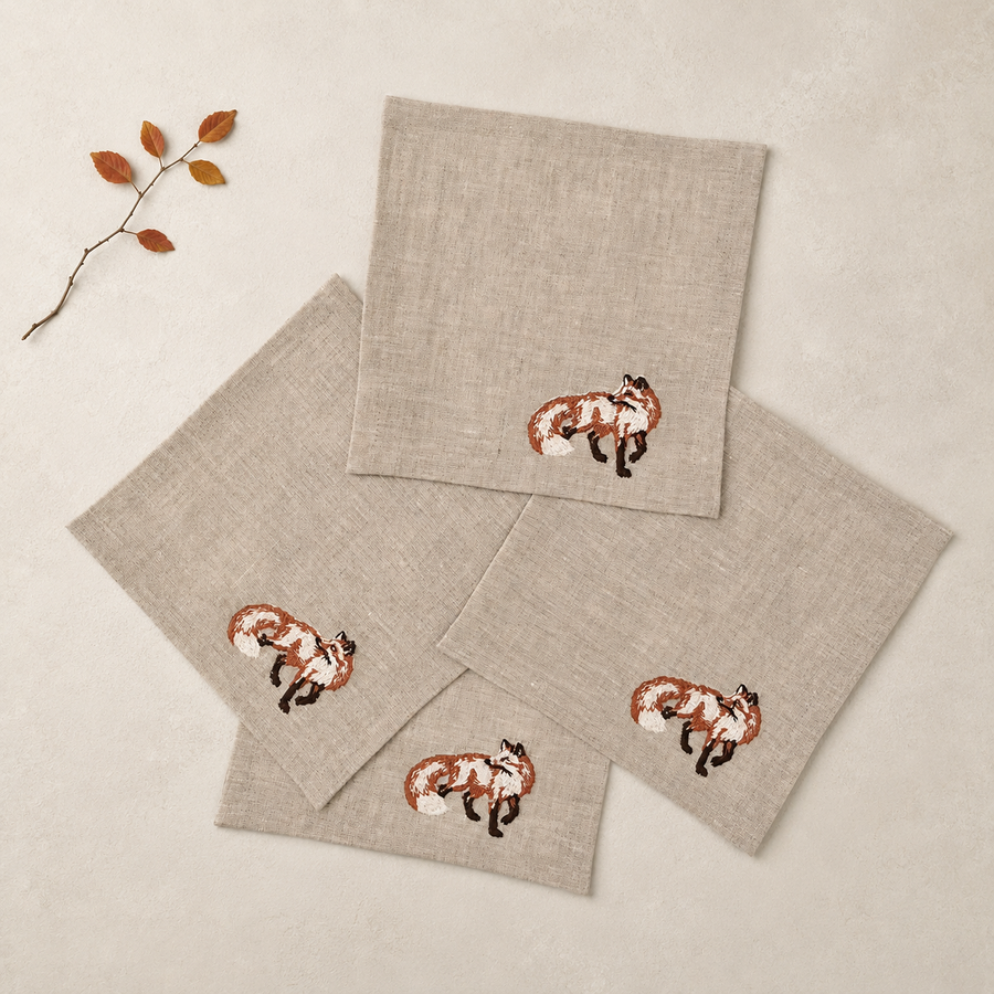 Four square linen napkins in a light beige color, each featuring a small embroidered fox motif in brown, orange, white, and black. The napkins are arranged in a flat-lay composition on a matching beige textured background with a small decorative dried leaf branch in the top-left corner.