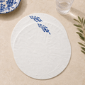 Two round white placemats with blue fish motifs, displayed on a beige stone surface with a glass of water and green leaves as props. MI RE LA