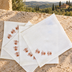 Four white linen napkins with embroidered brown bees, arranged on a rustic stone ledge with a scenic Tuscan landscape in the background. MI RE LA