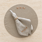 A folded light beige linen napkin tied with a rope ring, placed on a round matching beige placemat. The napkin and placemat both feature small embroidered bumblebee motifs near the top and bottom edges, set against a light beige stone surface. MI RE LA