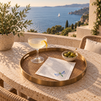 A round woven tray with a gold-toned rim sits on a stone table outdoors, styled with a cocktail glass, a napkin with a dragonfly motif, and a small bowl of olives. A terrace railing overlooks a blue sea and coastline. MI RE LA