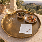 A round brass serving tray set on a stone table outdoors, styled with a coupe glass of white wine, a small bowl of olives, two slices of bruschetta, and a white embroidered napkin. A scenic vineyard landscape is visible in the background. MI RE LA