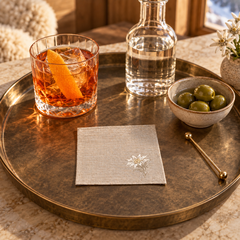 A styled tabletop scene with a round hammered metal tray holding a cocktail glass with an orange peel, a clear bottle of water, a small bowl of green olives, and a folded linen napkin with a white embroidered flower. The setting appears warm and rustic with wood and soft natural light. MI RE LA