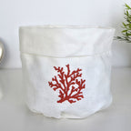 A white linen storage basket with a red coral embroidery on the front, set against a clean white background with a hint of green foliage on the right.