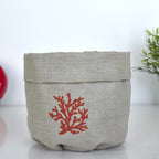 A light beige linen storage basket with a folded top edge and a red coral embroidery on the front, set against a white background with a hint of green foliage on the right.