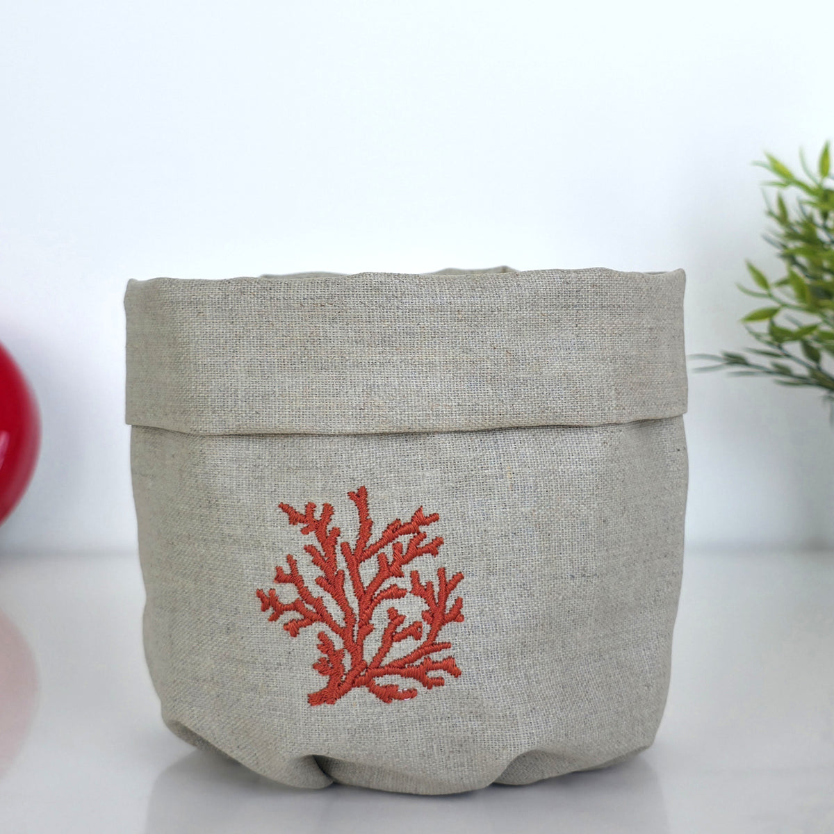 A light beige linen storage basket with a folded top edge and a red coral embroidery on the front, set against a white background with a hint of green foliage on the right.