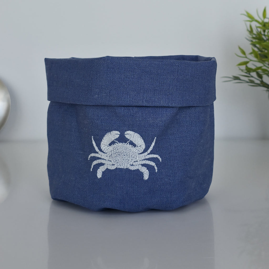A navy blue fabric storage basket with a white embroidered crab design on the front, set on a reflective gray surface with a soft-focus background.