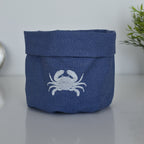 A navy blue fabric storage basket with a white embroidered crab design on the front, set on a reflective gray surface with a soft-focus background.