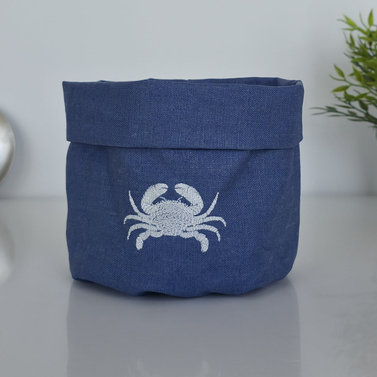 A navy blue fabric storage basket with a white embroidered crab design on the front, set on a reflective gray surface with a soft-focus background.