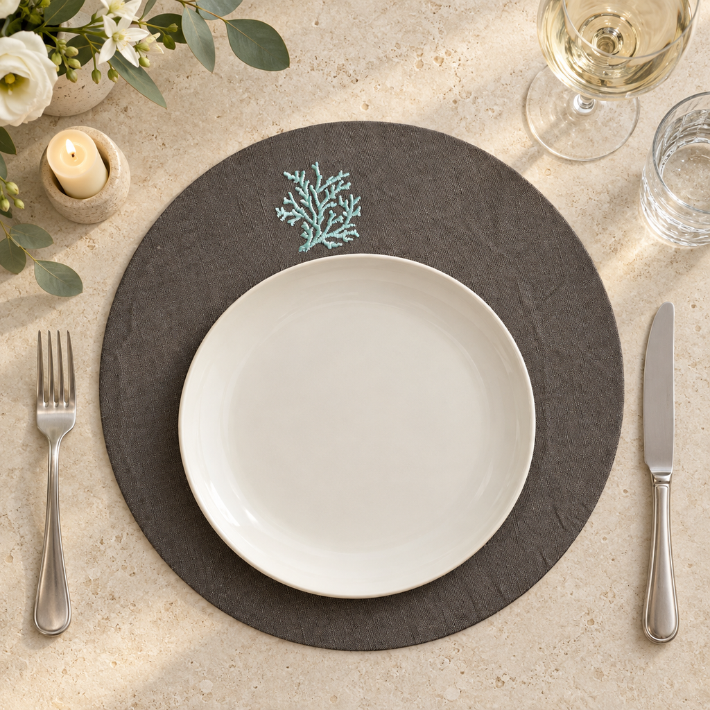 A styled table setting featuring a round dark gray placemat with a light blue coral motif, layered with a white ceramic plate, silver fork, and knife on a beige stone surface. MI RE LA