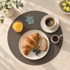 A styled breakfast scene on a round dark gray placemat over a light beige table. A white plate holds a golden croissant and a slice of bread with blueberries, alongside a cup of coffee on a saucer, a glass of orange juice, a small jam dish, and a vase of white flowers. MI RE LA