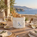 A offwhite coated linen bread basket with a blue dragonfly embroidery sits on a wooden outdoor dining table, filled with breadsticks and round bread, with a scenic sea view in the background.