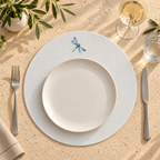 A styled table setting featuring a round white plate centered on a light off whiteplacemat with a small blue dragonfly illustration near the top. A silver fork is on the left and a knife on the right, all arranged on a textured beige tabletop with olive branches, a glass of white wine, and a glass of water nearby. MI RE LA