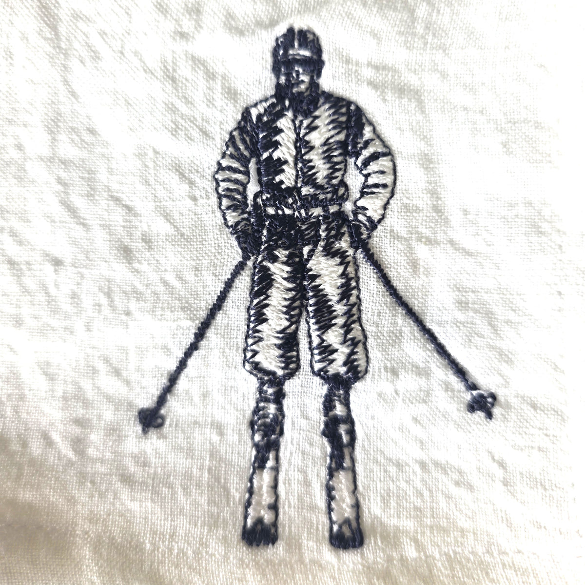 A close-up of a white textured fabric, likely a napkin or decorative cloth, featuring a black embroidered illustration of a skier in a vintage ski suit holding ski poles.