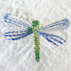A close-up of a hand-embroidered dragonfly on textured white fabric, featuring blue and white striped wings and a green body. MI RE LA